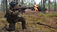 Ein Soldat mit einem Maschinengewehr kniet im Wald und schießt. An der Mündung leuchtet Feuer auf.