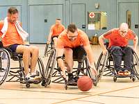 Drei Sportler im Rollstuhl kämpfen um einen auf dem Boden rollenden Basketball.
