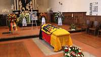 In einer Kirche steht ein Soldaten-Foto zwischen Blumenkränzen. Davor steht ein Sarg umhüllt mit der Bundesdienstflagge.