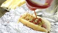 Ein Hot-Dog liegt auf Alufolie und wird mit Ketchup gewürzt. Im Hintergrund liegen zwei Wraps auf der Kochplatte.