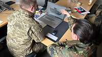 Ein deutscher und ein ukrainischen Soldat besprechen etwas an einem aufgeklappten Laptop, auf dem Panzer zu sehen ist.