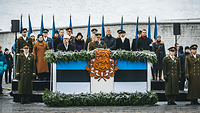 Auf einer Tribüne, die mit einem Wappen und Flaggen Estlands geschmückt ist, stehen zahlreiche Politiker und Militärs.