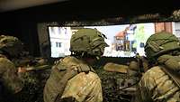 Soldaten in Gefechtsausrüstung sitzen in einem Simulator und haben ein Maschinengewehr im Anschlag. 