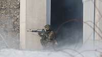 Ein Soldat kniet neben einem Hauseingang mit seiner Waffe im Anschlag und sichert die Hausecke ab.