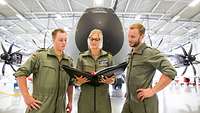 Drei Soldaten in Fliegerkombi besprechen sich vor einem militärischen Transportflugzeug in einer Wartungshalle
