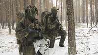 Zwei getarnte Soldaten knien im verschneiten Waldboden.
