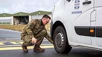 Ein Soldat kniet neben einem Van, während im Hintergrund eine Flugzeughalle steht 