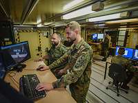Zwei Soldaten arbeiten im Stehen an einem Computer, auf dem Luftbildaufnahmen zu sehen sind.