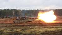 Mehrere Panzer befinden sich auf einem Gelände, aus einem Kanonenrohr kommt ein großer Feuerball