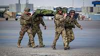 Soldaten aus mehreren Nationen haben einen Verwundeten auf einer Trage und marschieren entlang eines Flugfeldes