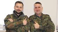 Zwei Soldaten posieren mit verschränkten Armen nebeneinander vor einer Wand und lächeln in die Kamera. 
