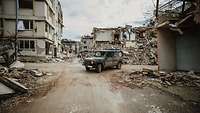 Ein Militärfahrzeug mit rotem Kreuz steht auf einer unbefestigten Straße vor Trümmern und zerstörten Gebäuden.