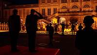 Mehrere Soldaten stehen grüßend nachts beim Appell auf einem Schlossplatz mit Fackeln.