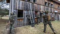 Vor einem Holzhaus stehen Soldaten mit Leiter. Einer wirft eine Handgranate in den ersten Stock.