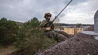 Ein Soldat in Flecktarnuniform hängt mit einem Seil an einer Häuserkante.