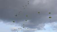 Aus einem Flugzeug springen Fallschirmjäger ab, zahlreiche grüne Schirme segeln unter dem Himmel.