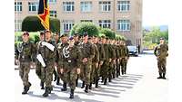 Eine Gruppe Soldaten marschiert in einer Kaserne. Vorneweg die Ehrenformation mit Deutschland-Fahne 