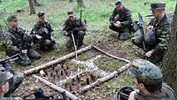 Gruppe Soldaten im Wald vor einem Modell der Umgebung aus Stöcken. Ein Soldat zeigt ihre Übungsräume.