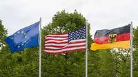 Die deutsche, die amerikanische und die europäische Flagge wehen zusammen an Fahnenmasten.