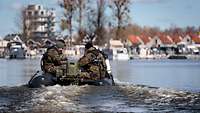 Mehrere Soldaten in Flecktarn sitzen in einem schwarzen Gummiboot und fahren auf einem Gewässer.