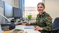 Eine Soldatin sitzt an einem Schreibtisch blickt in die Kamera. Sie hat ein Buch aufgeschlagen und lächelt.