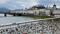 Ein Brückengeländer mit vielen Schlössern im Hintergrund die Sicht auf Salzburg. 