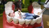 Ein Junge liegt auf den Zähnen eines übergroßen Plastikgebisses, dessen Zahn gerade von einem übergroßen Bohrer bearbeitet wird.