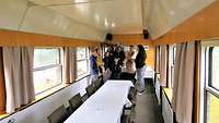 Eine Besuchergruppe besichtgt einen ehemaligen Konferenzwagen der Reichsbahn, der mit Tischen und Stühlen ausgestattet ist. 