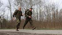 Zwei Soldaten in Flecktarnuniform und mit Rucksäcken rennen lächelnd auf einem Waldweg.
