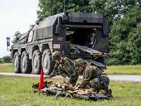 Zwei Soldaten versorgen eine verletzte Person, im Hintergrund steht ein Radpanzer mit rotem Kreuz.