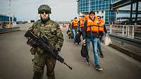 Ein bewaffneter Soldat geleitet mehrere Personen mit Schwimmwesten im Hafen.