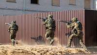 Drei Soldaten mit Maschinengewehren rennen an einer Mauer entlang.