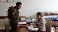 Die rechts sitzende Sekretärin schaut in einem Büro in Unterlagen. Ein Soldat beugt sich von links leicht über den Schreibtisch.