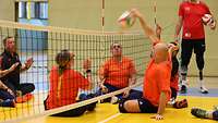 Zwei Teams spielen gegeneinander Sitzvolleyball, eine Person wirft dabei den Ball über das Netz