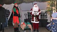 Der Weihnachtsmann und eine Elfe stehen stehen nebeneinander, ein Bollerwagen steht dazwischen. 