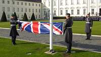 Zwei Soldaten falten eine Flagge wärend die anderen Soldaten salutieren mit Blick zur Flagge. 