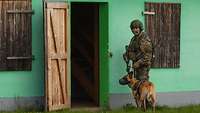 Ein Soldat steht mit einem Schäferhund neben der geöffneten Holztür eines grünen Hauses.