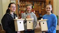 Drei Schülerinnen stehen in einer Tischlerei der Bundeswehr und zeigen neu gebaute Vogelhäuschen