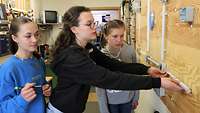 Drei Mädchen vermessen mit einem Zollstock in einer Werkstatt einen Schaltkreis an einer Montagewand