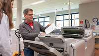 Ein Mitarbeiter zeigt einem Mädchen ein Druckerzeugnis vor einer Maschine