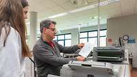 In einem Raum mit Maschinen zeigt ein Mitarbeiter der Schülerin das Druckergebnis