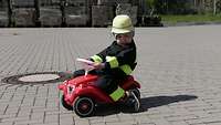 Ein Kind sitzt in einer Feuerwehruniform auf einem roten Bobby-Car. 