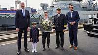 Fünf Personen stehen vor einem Kriegsschiff. Eine Frau hält einen Blumenstrauß. 