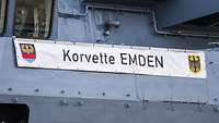 Eine Plane mit dem Wappen der Stadt Emden, der BRD und den Worten "Korvette Emden" hängt am Schiff.