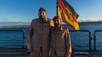 Zwei Soldaten stehen nebeneinander an Bord eines Schiffes, hinter ihnen weht die deutsche Flagge. 