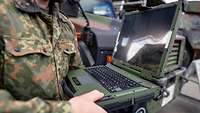 Ein Soldat steht vor Fahrzeugen und hält ein Laptop in einer olivfarbenen Metallhülle.