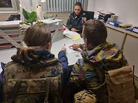 Eine Frau und ein Mann in Flecktarnuniform sprechen, in einem Büro sitzend, mit einer Polizistin.