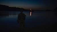 Eine Meeresbucht in der späten Abenddämmerung; im Vordergrund ein Soldat in grüner Flecktarnuniform.