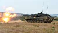 Ein Kampfpanzer Leopard 2 schießt scharf im Gelände. Aus dem Kanonenrohr tritt Feuer aus.