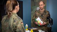 Eine Soldatin spricht in einem Studio mit einem Soldaten, der eine Tasche in seinen Händen hält.